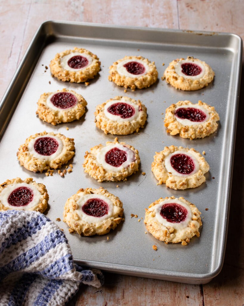 Cooked raspberry cream cheese thumbprint cookies on a baking sheet with a blue and white striped towel.