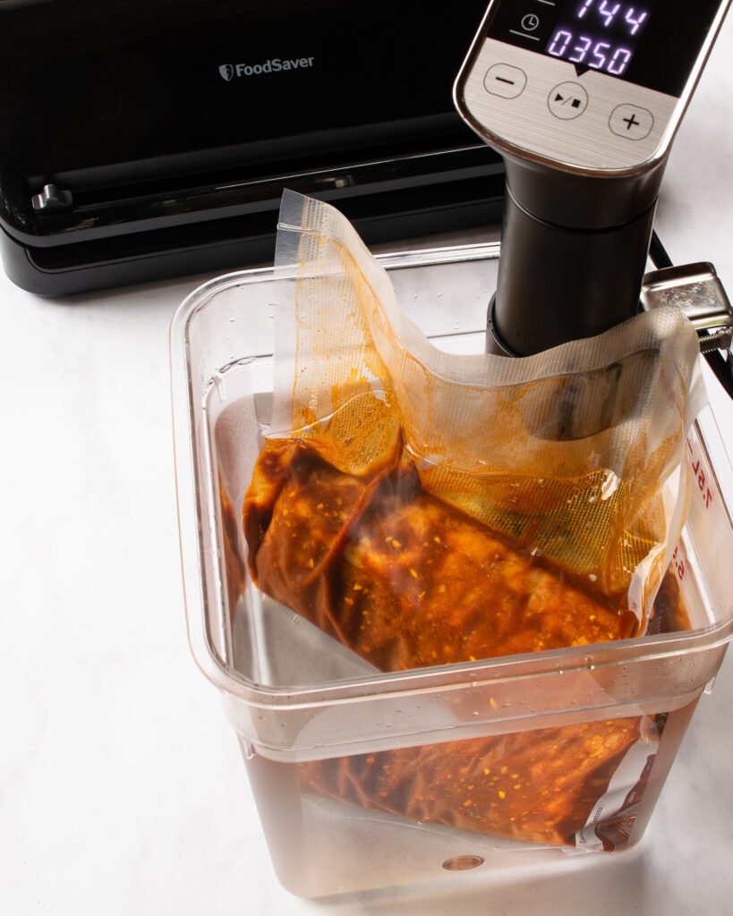 A sous vide water bath cooking a pork loin with a food saver vacuum sealer in the background.