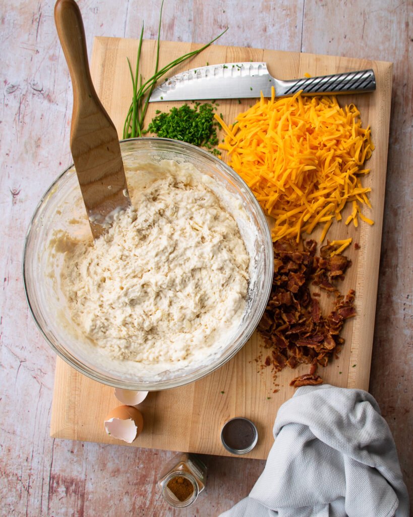 Ingredients on a wooden board - aa bowl of batter, grated Cheddar cheese, cooked bacon and chopped chives.