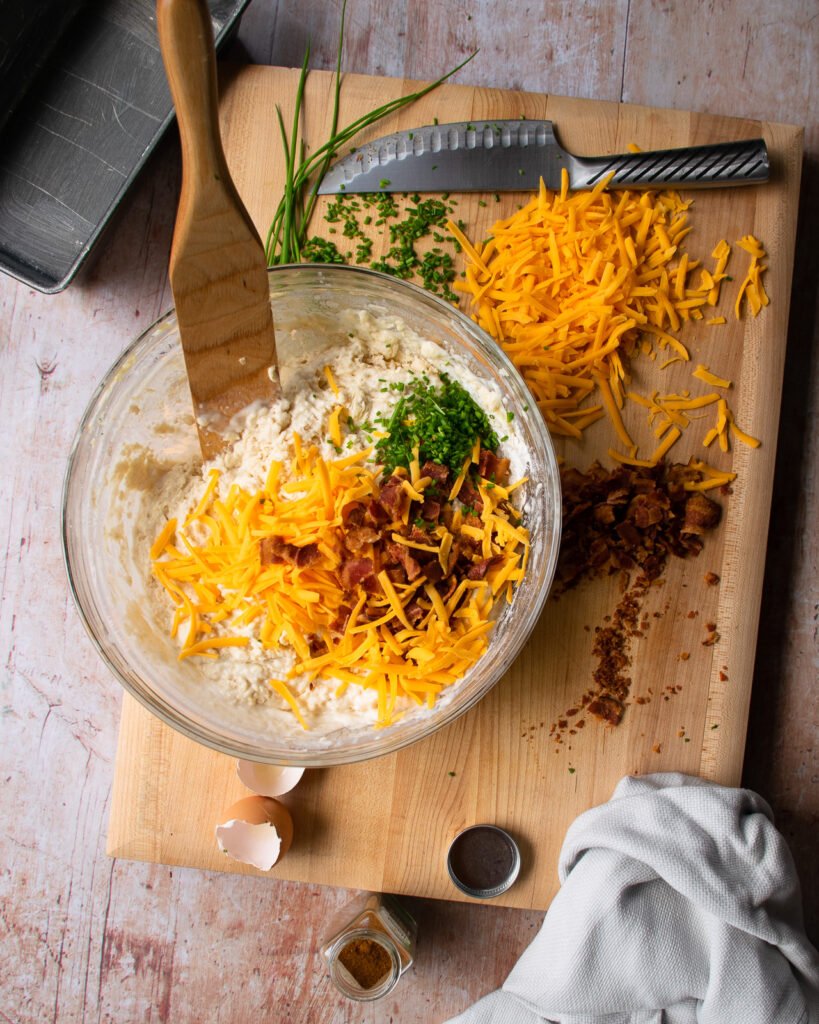 Stirring cheese, bacon and chives into a batter of buttermilk biscuit bread. A loaf pan in the background.