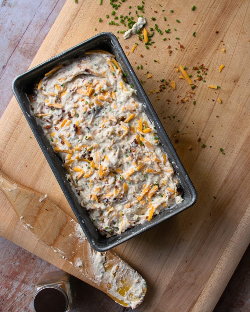 Buttermilk biscuit bread batter in a loaf pan, ready to go into the oven, on a wooden cutting board.