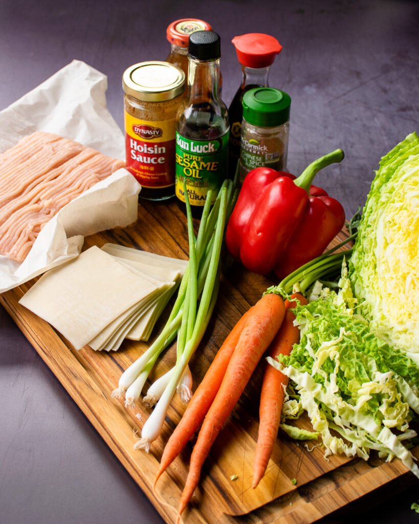 Ingredients on a cutting board - cabbage, carrots, green onions, wonton wrappers, soy sauce, hoisin sauce, sesame oil, red pepper and chicken.