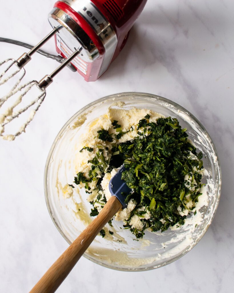 A glass bowl with cream cheese, butter and spinach in it, a spatula sticking out of the bowl and a hand mixer resting near by.