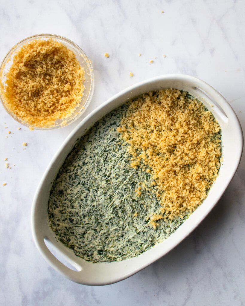 Creamed spinach casserole being assembled in a white ceramic oval dish and breadcrumbs on top of half of it.