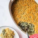 Creamed Spinach Casserole in a white oval ceramic dish with a spoonful removed and on a side plate.