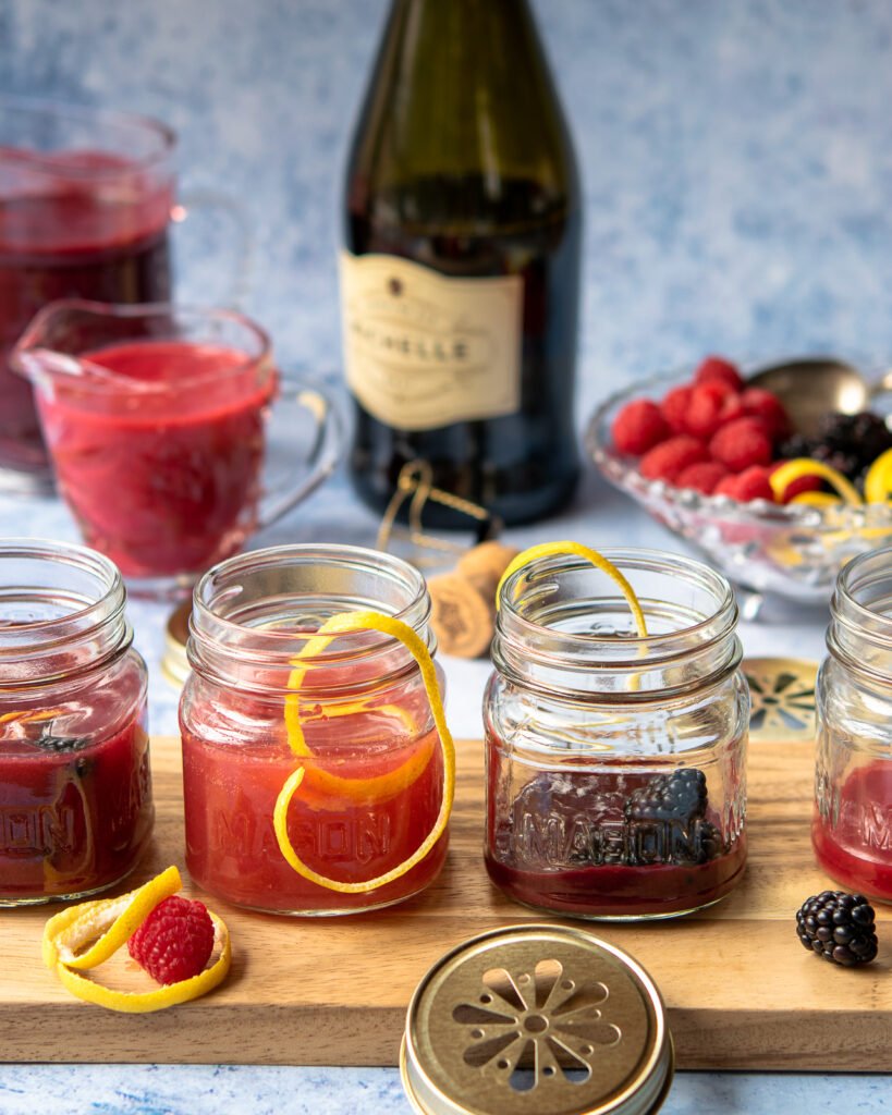 Mason jars with jam bellinis lined up on a piece of wood with prosecco in the background.