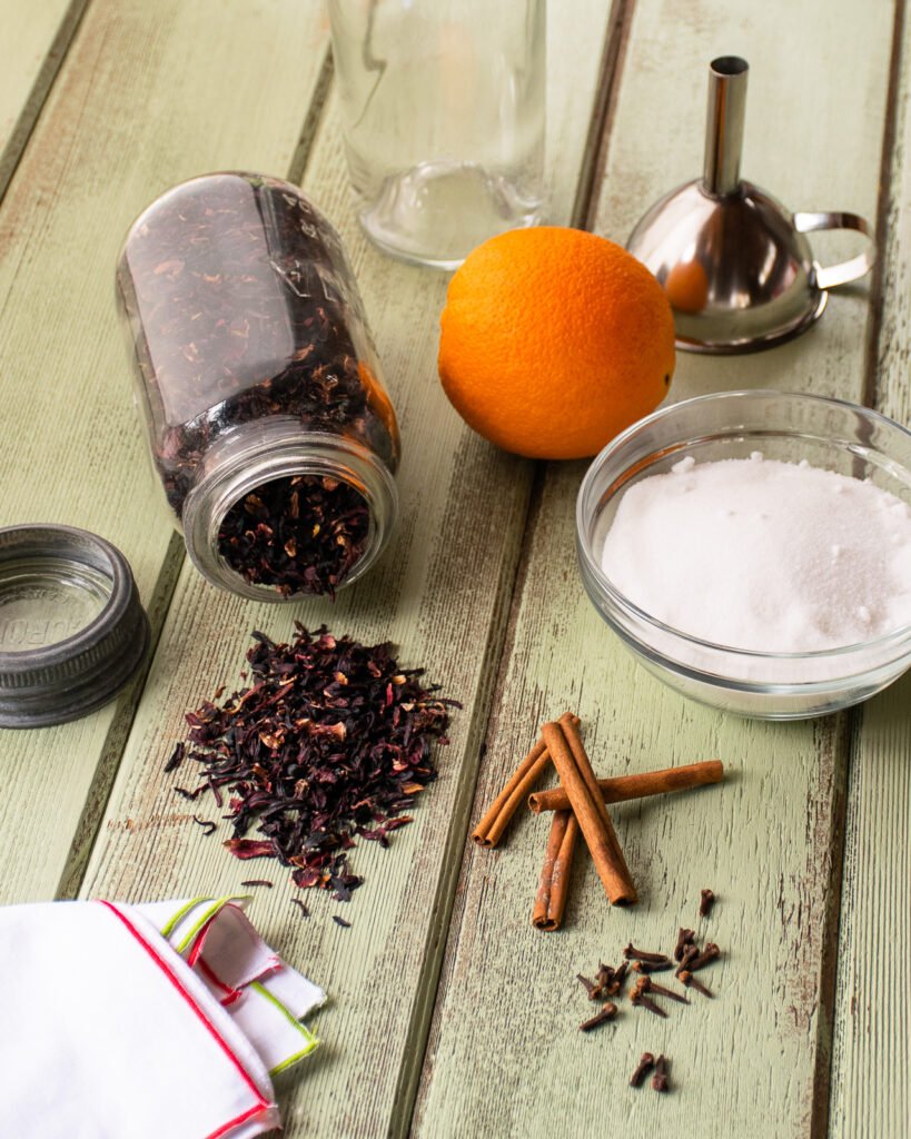 Ingredients and equipment to make sorrel on a green table - an orange, cinnamon sticks, sugar, cloves, hibiscus flowers and a funnel.
