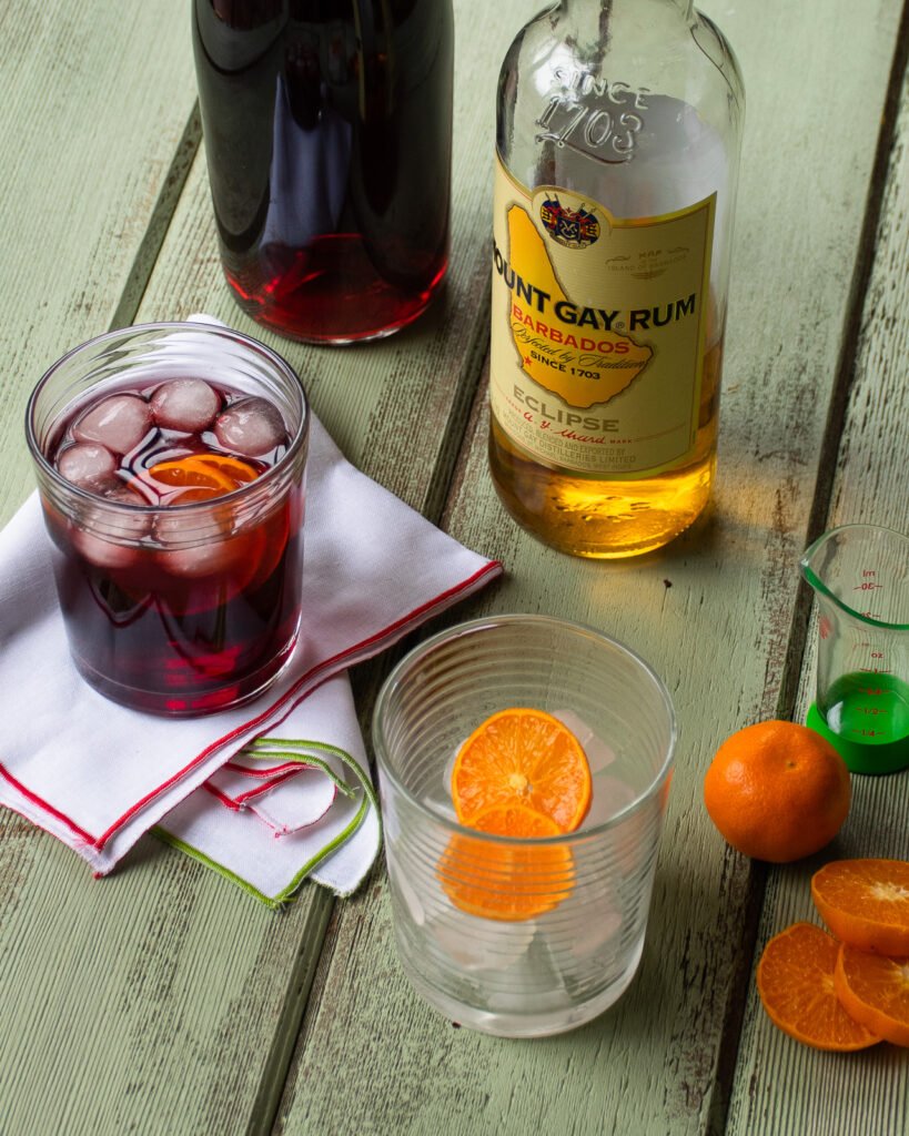 An empty glass with a mandarin slice, a full glass with sorrel cocktail, a bottle of sorrel and a bottle of rum on a green table.
