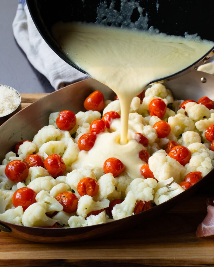 Cauliflower and roasted tomatoes in a gratin dish with alfredo sauce being poured on top.