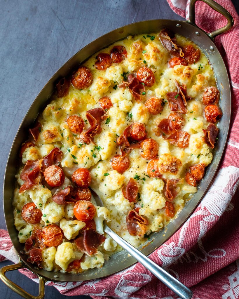 Cauliflower Alfredo Bake in an oval gratin dish on a counter with a red towel wrapped around the pan and a spoon sticking out.