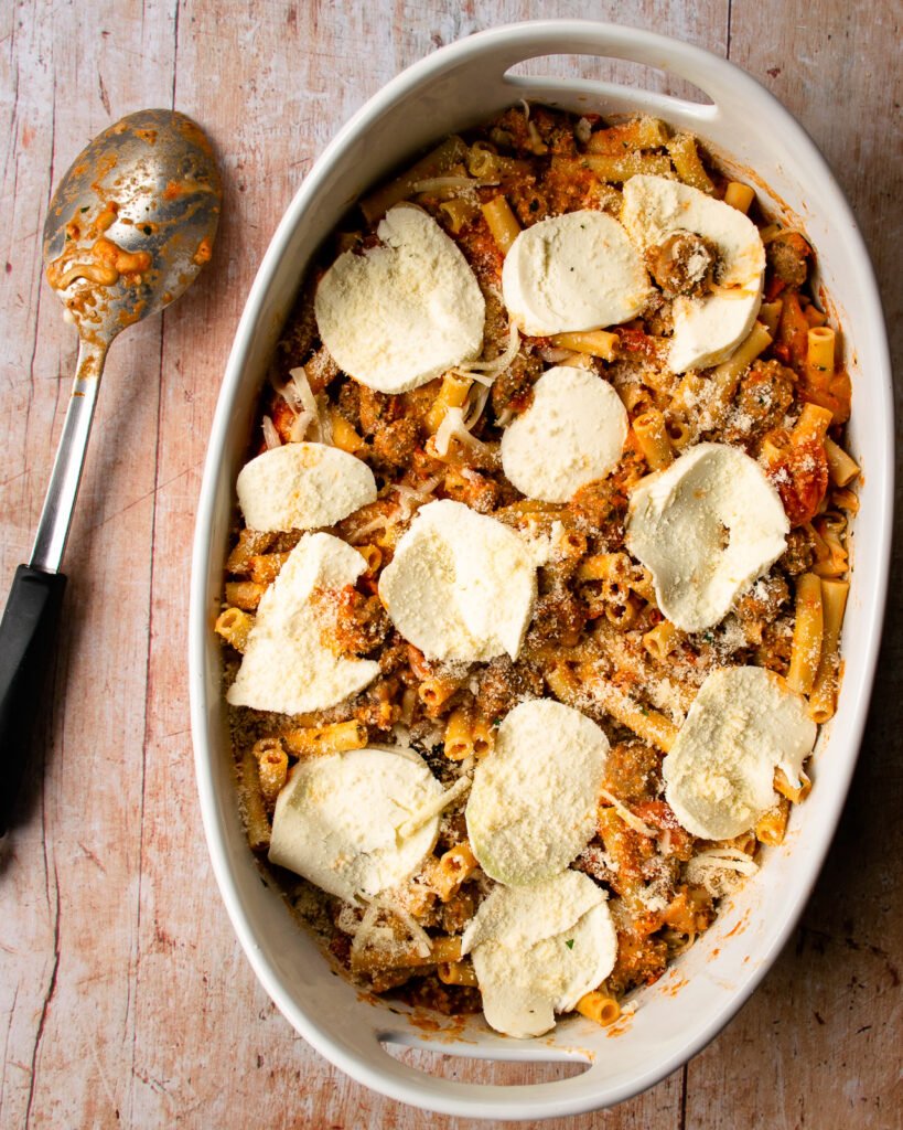 Ziti al forno assembled in a white oval ceramic dish and a spoon alongside.