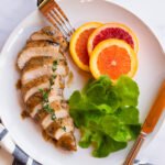 Marinated chicken breast on a white plate with a few salad greens and slices of citrus.