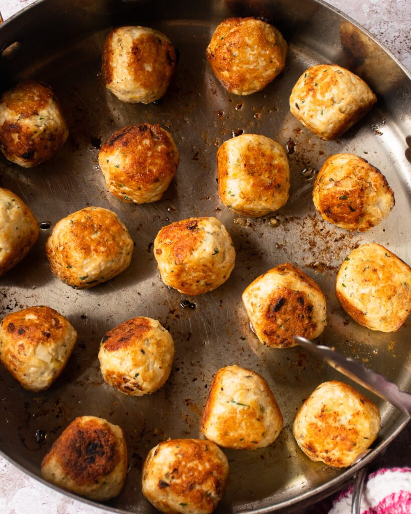 Chicken meatballs cooking in a saute pan with tongs resting on the side.