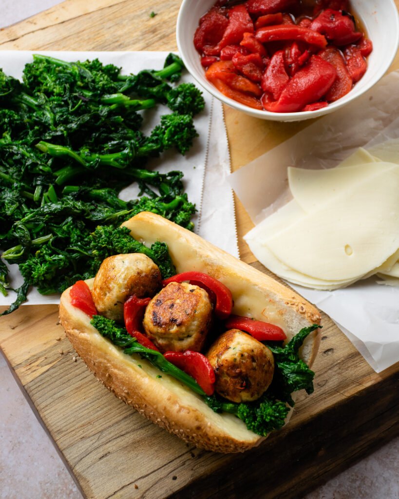 Building Italian Chicken Meatball Grinders - one on a cutting board with a bowl of roasted red pepper, some slices of provolone cheese and cooked broccoli rabe next to it.