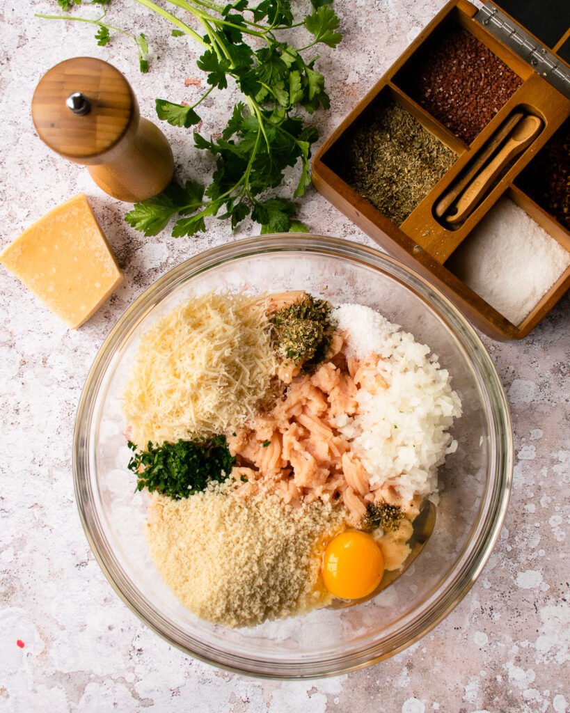 Ingredients for meatballs in a glass bowl on a counter with parmesan cheese, a pepper grinder, some parsley and spices near by.