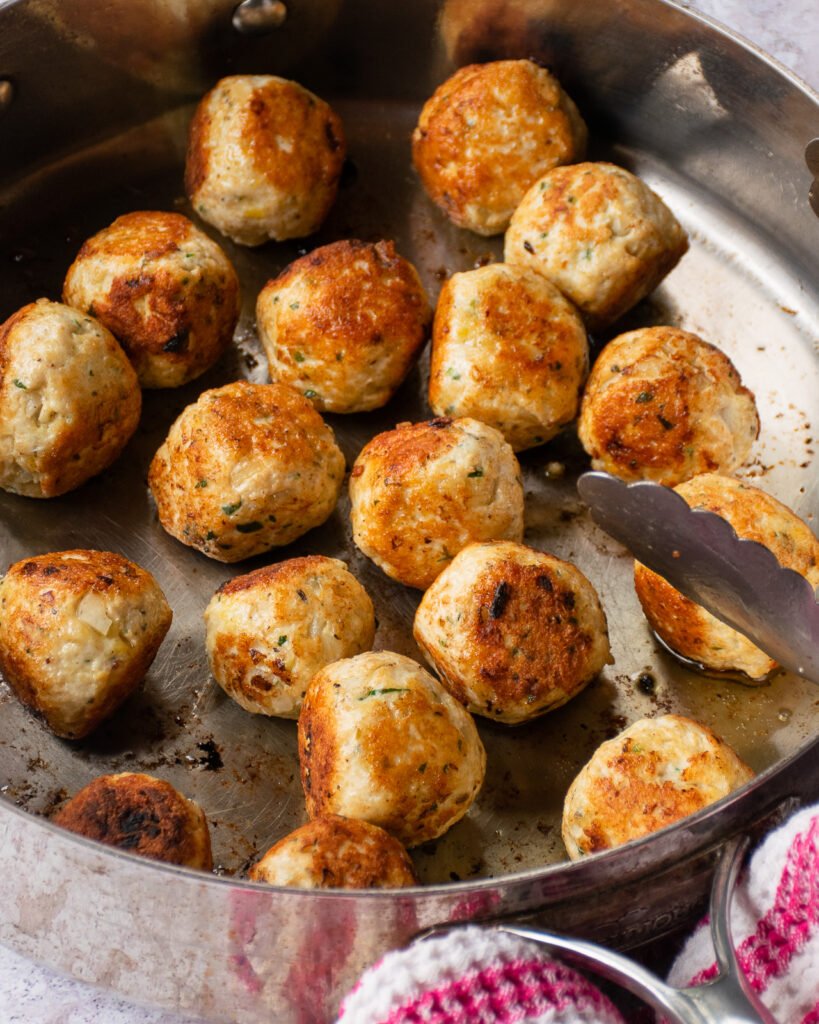Chicken meatballs in a saute pan with a pair of tongs resting on the side.