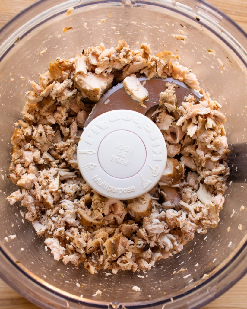 Looking straight down into a food processor with chicken chopped up inside.