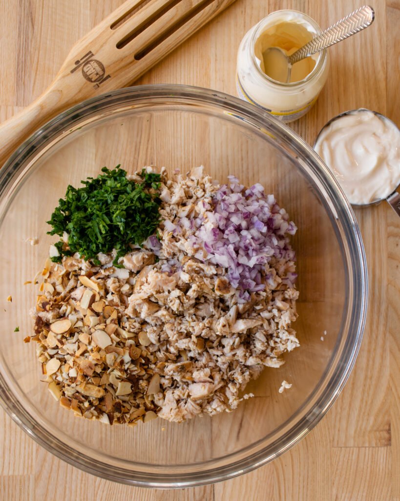 A glass bowl with chopped chicken, red onion, toasted almonds and parsley, next to a bottle of mayonnaise and a wooden spoon.