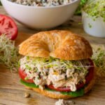 A chicken salad croissant sandwich on a wooden cutting board with a bowl of chicken salad in the background.