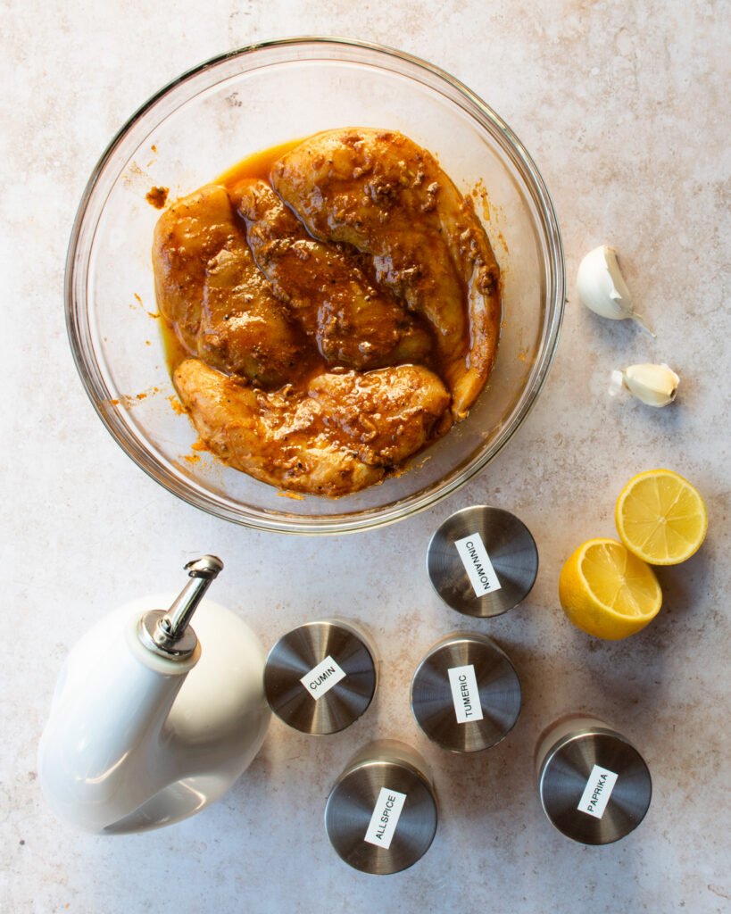 Chicken marinating in a bowl with ingredients around the bowl - spices, lemon, garlic and oil.