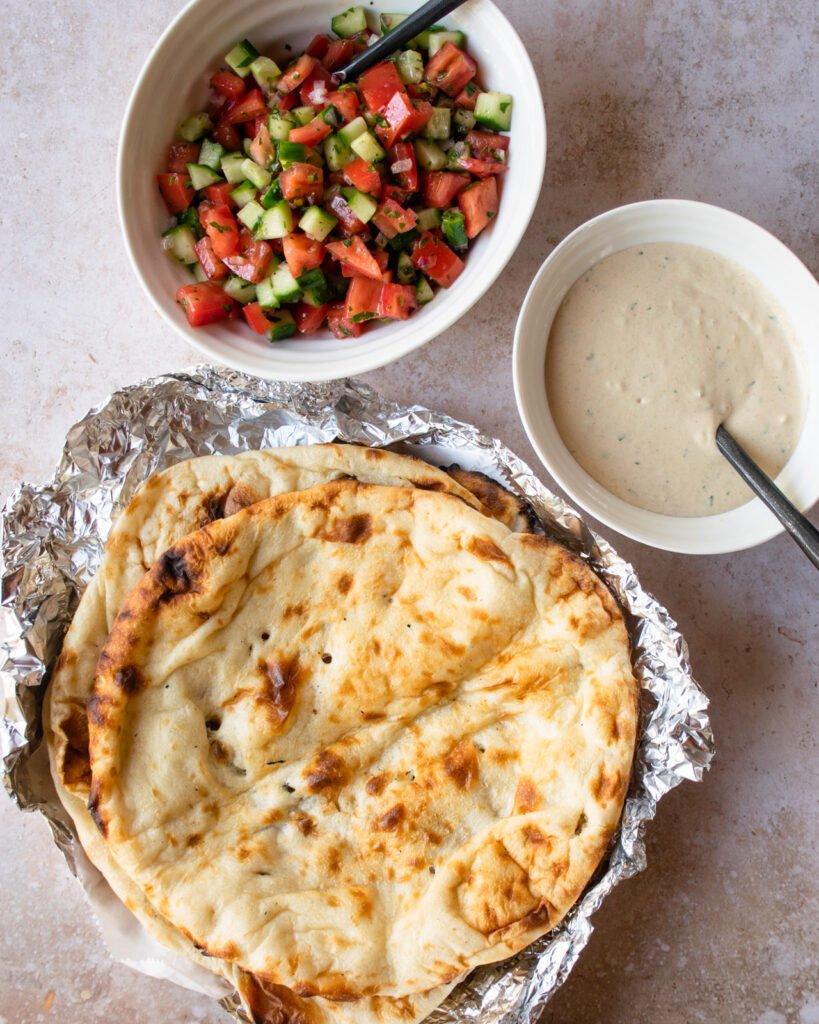 Baked naan bread in foil, tahini sauce in a white bowl and tomato cucumber salad in a white bowl all on a tan countertop.