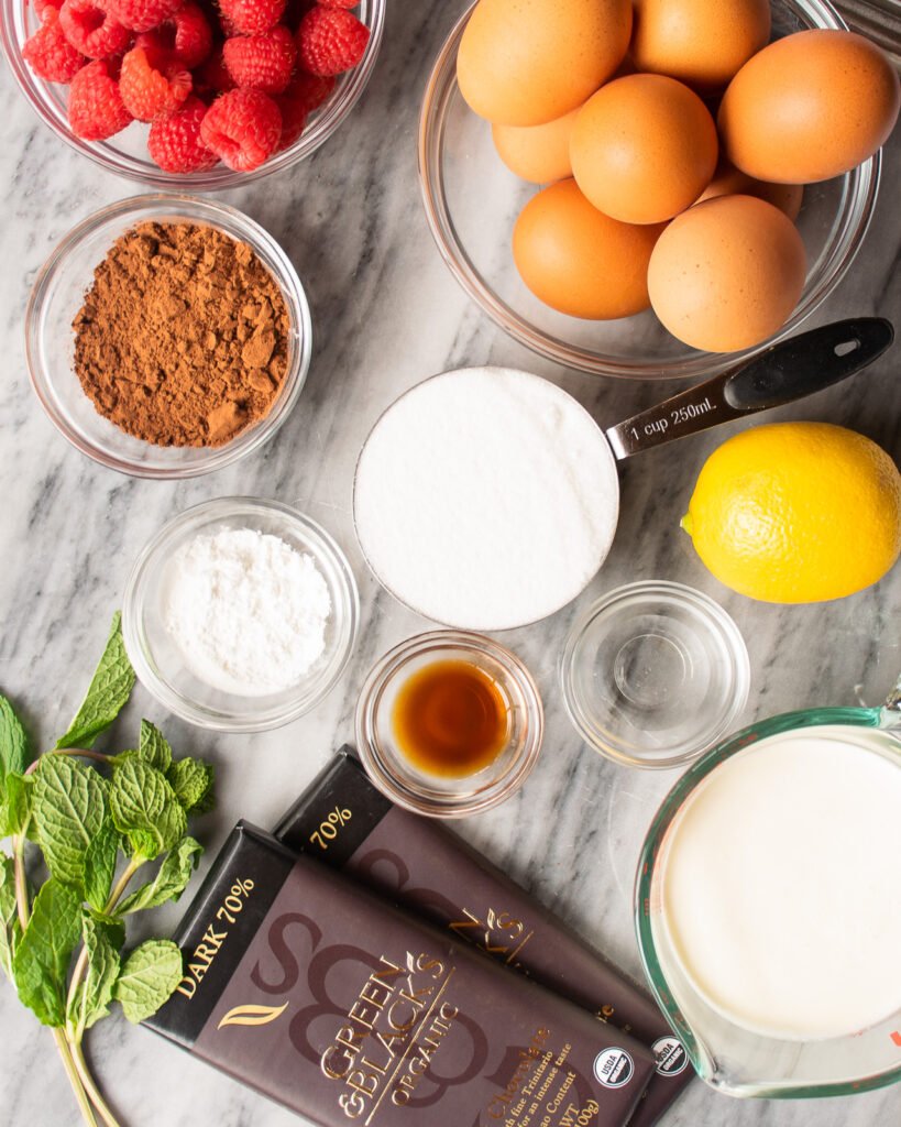 Ingredients on a marble counter - chocolate, mint, sugar, lemon, vanilla, vinegar, cornstarch, cocoa powder, eggs and raspberries.