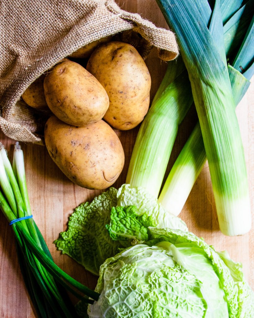 A burlap basket of potatoes, a head of cabbage, some scallions and leeks on a wooden counter.