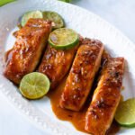 Four fillets of honey soy salmon on a white plate with lime slices and a green napkin.