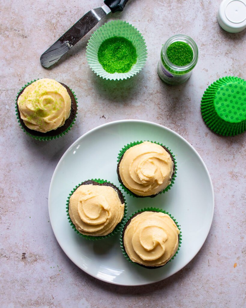 Irish Coffee Cupcakes with a plain swirl design on a plate with green cupcake liners and green sprinkles on the counter.