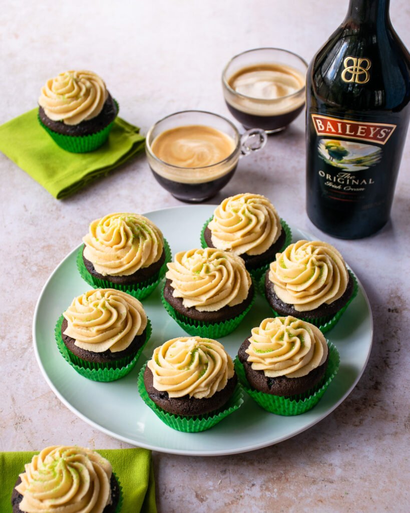 Irish Coffee Cupcakes on a plate with frosting piped on with a star tip and a bottle of Bailey's and two coffees in the background.