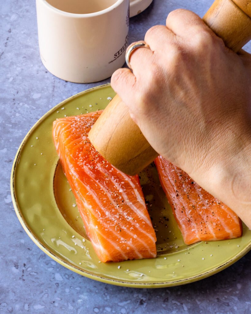 Hands grinding pepper onto salmon fillets on a green plate.