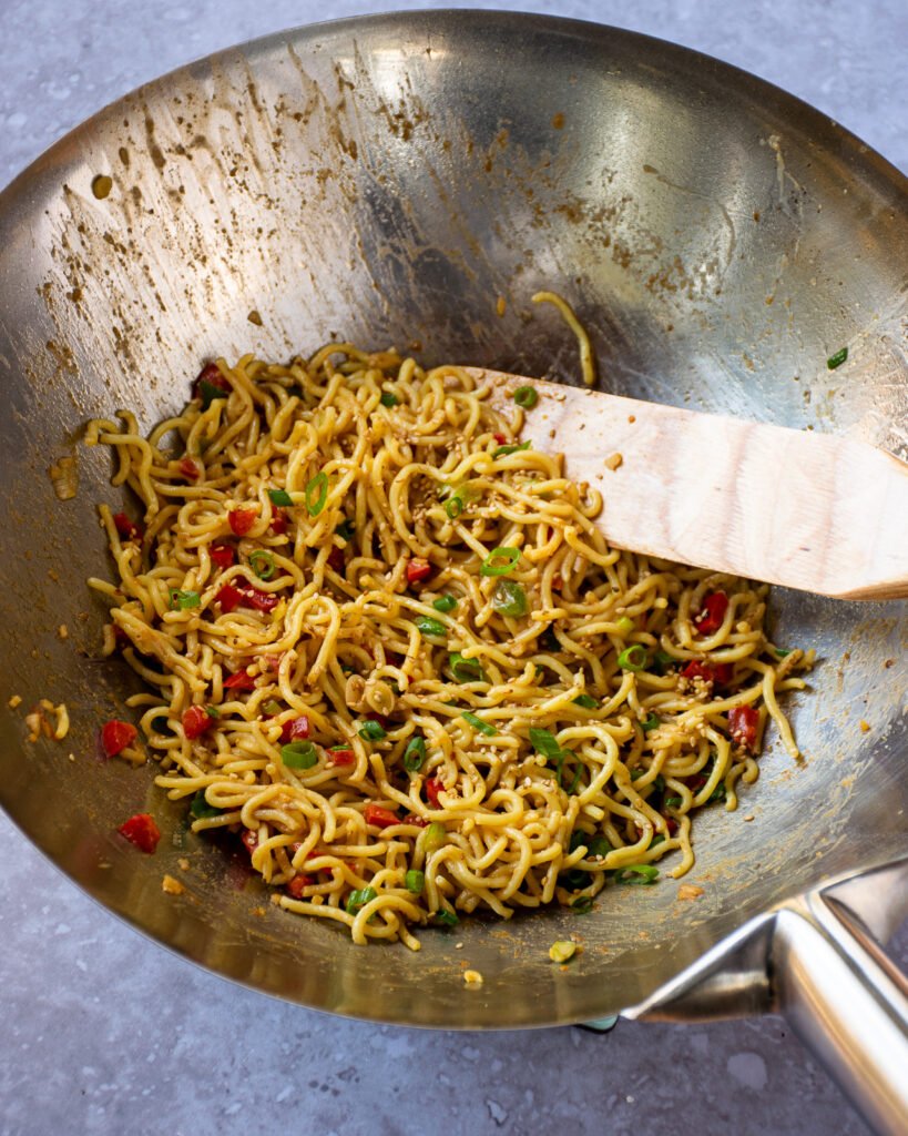 Sesame Noodles stir-frying in a silver wok with a wooden spoon.