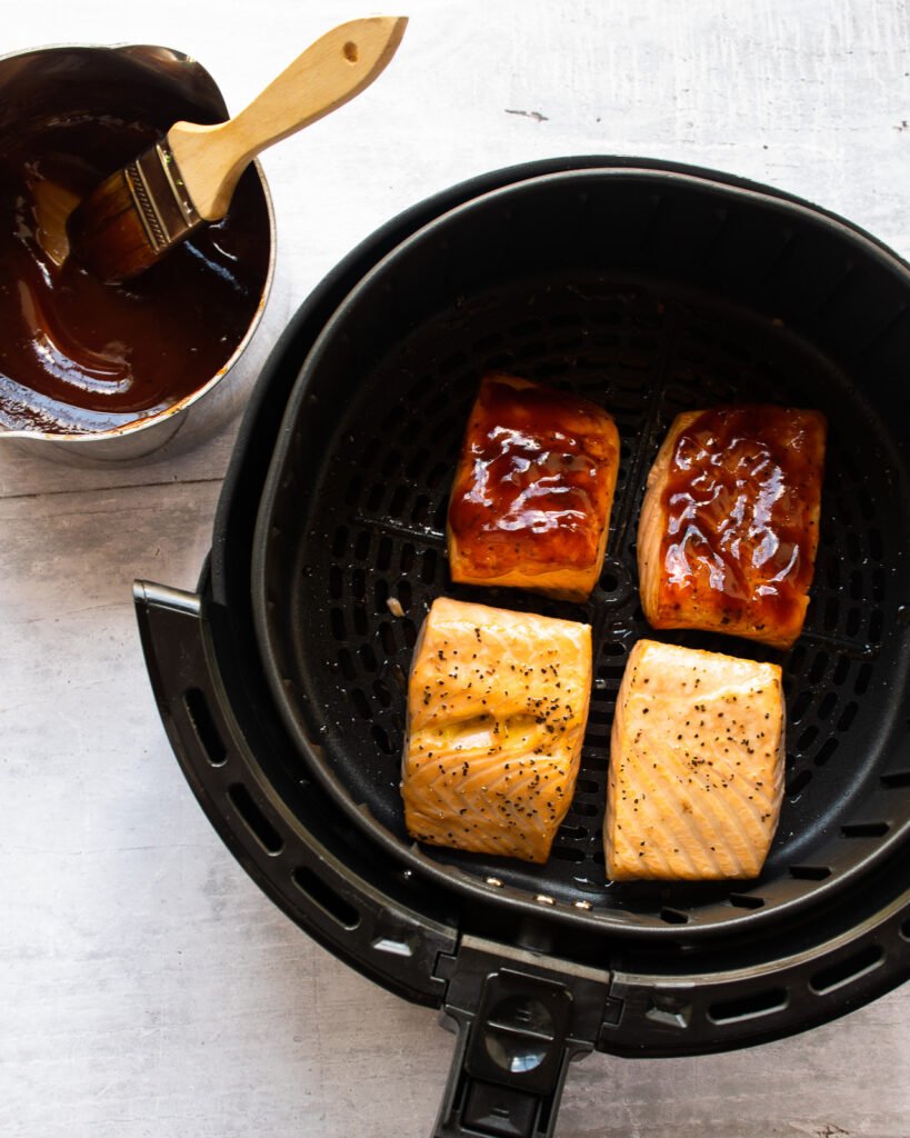 Four salmon fillets in an air fryer basket with a saucepot of BBQ sauce and basting brush next to it.