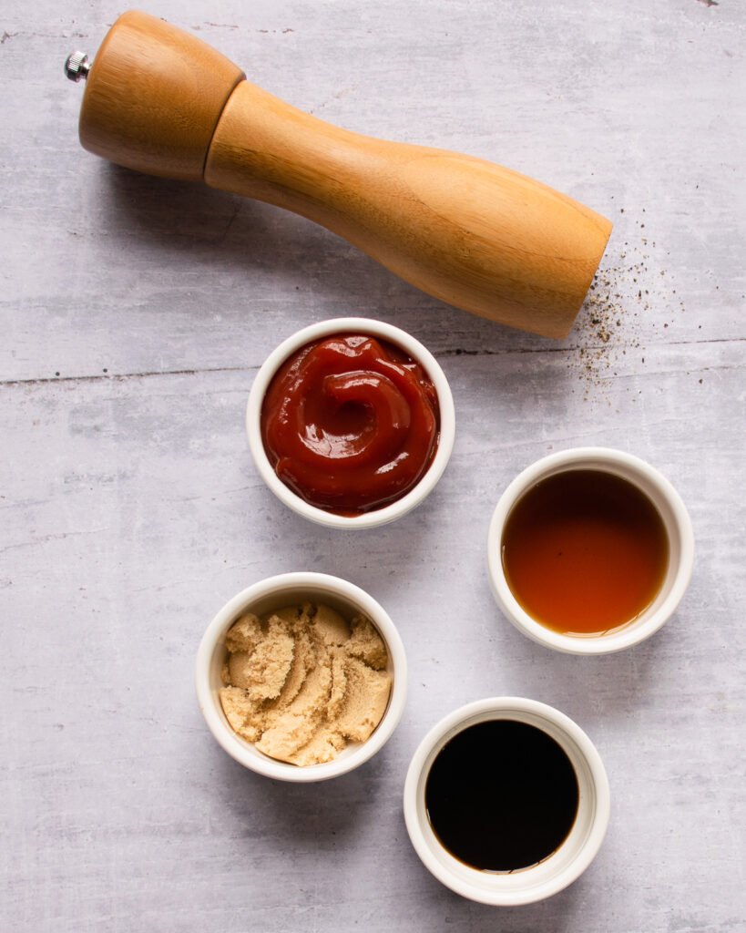 Ingredients on a wooden counter - ketchup, dry mustard, white balsamic vinegar, molasses and a peppermill.