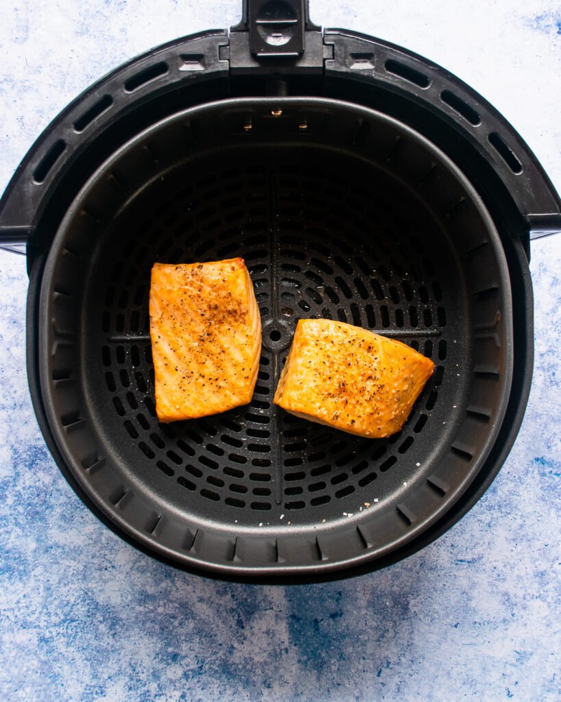 Two cooked fillets of salmon in an air fryer basket with a blue background.