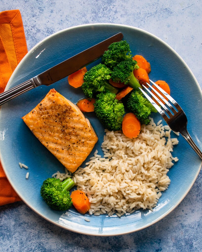 A blue plate with air-fried salmon, rice and vegetables, knife and fork on it, with an orange napkin beside it.