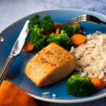 A blue plate with a fillet of air-fried salmon, rice and vegetables on a blue counter.