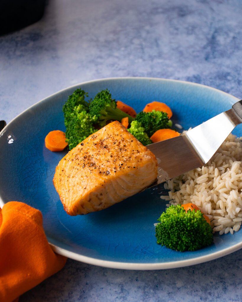 A spatula placing a fillet of salmon on a blue plate with rice and vegetables.