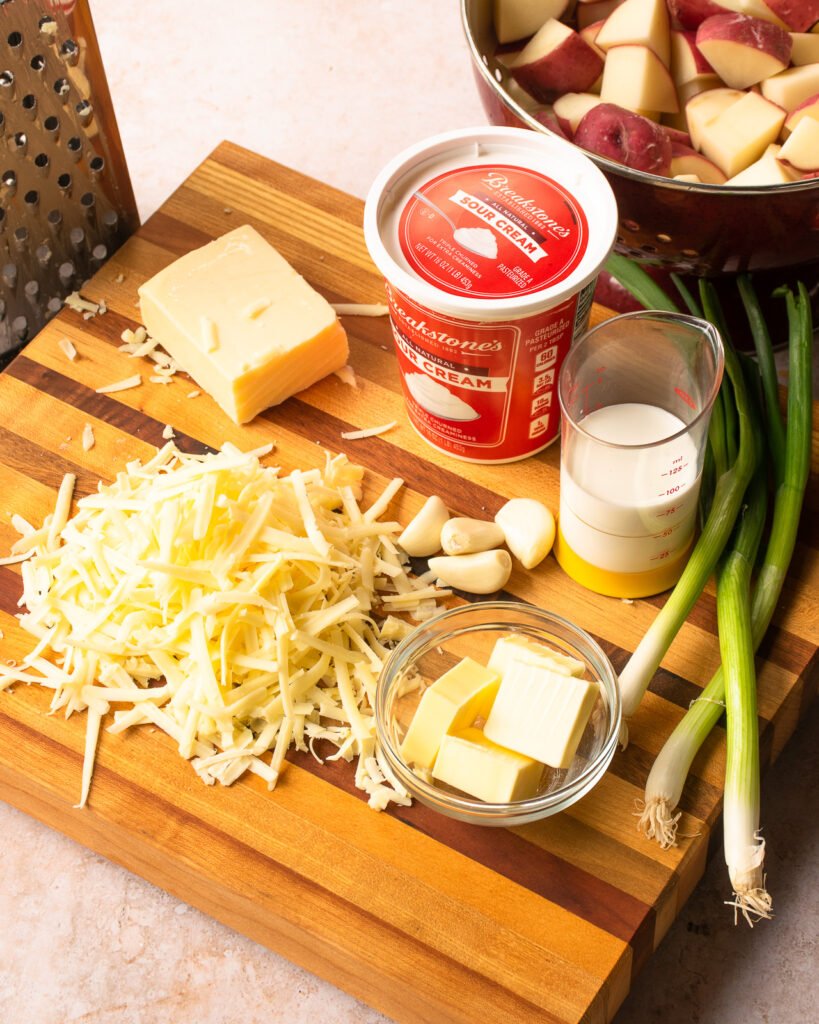 Ingredients on a cutting board - grated Cheddar cheese, butter, sour cream, heavy cream, scallions - next to a grater and a colander with chunks of red potatoes.