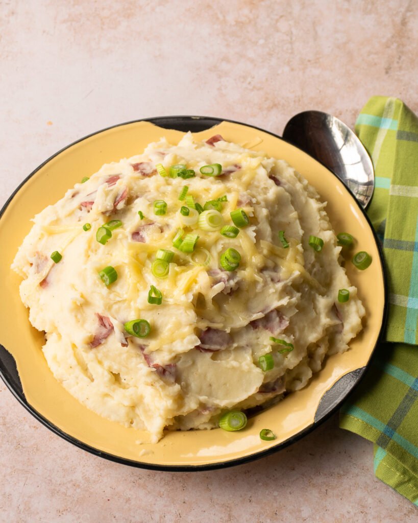 Cheddar garlic smashed potatoes with scallions on top in a rustic yellow bowl and a green plaid napkin beside it.