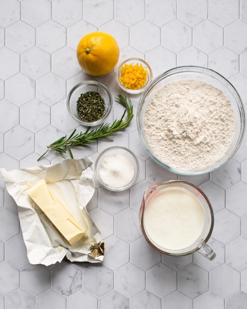 Ingredients on a counter - flour, buttermilk, lemon, rosemary, baking powder and butter.