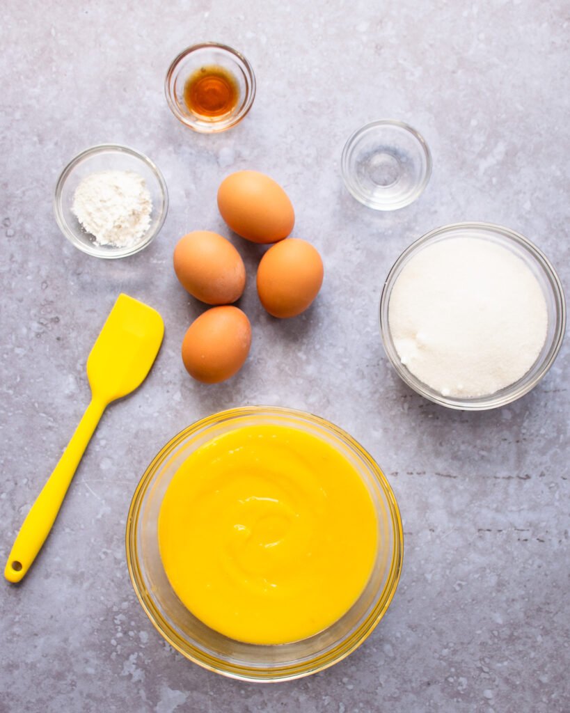 Lemon curd, eggs, sugar, vanilla, vinegar and cornstarch on a countertop with a yellow spatula.