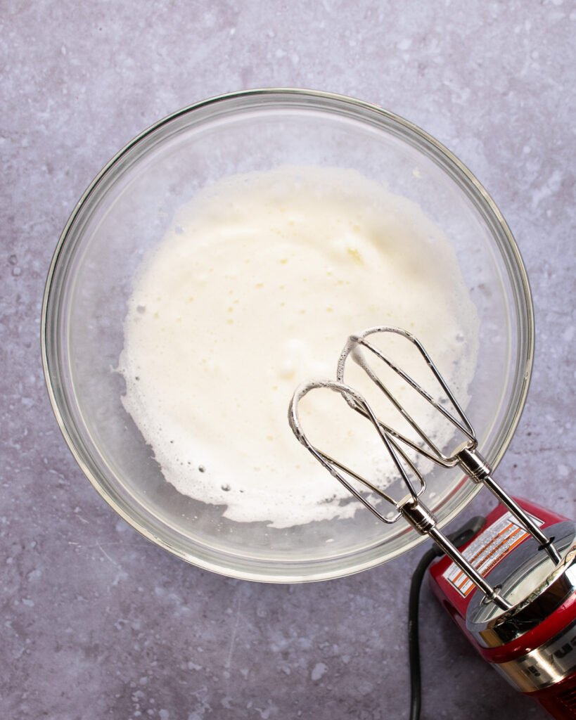 A bowl of egg whites partially whipped in a glass bowl with a hand mixer next to it.