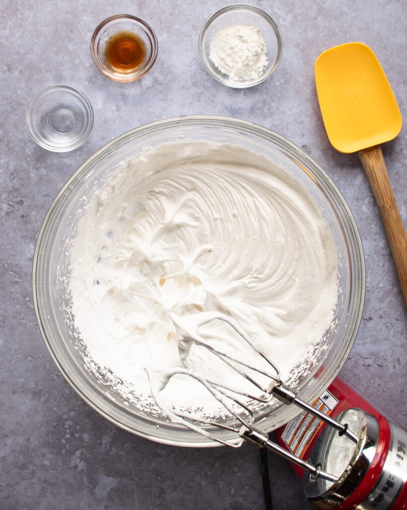 Meringue in a glass bowl with vanilla, vinegar and cornstarch on a countertop.