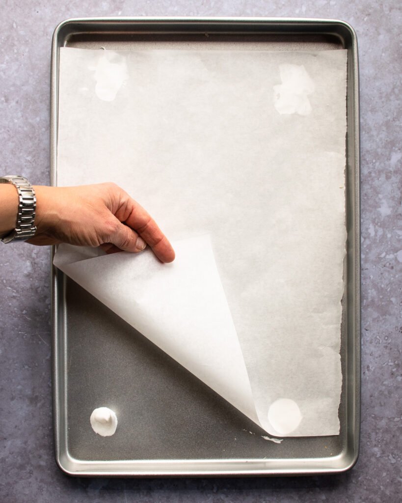 A hand holding parchment paper up on a baking sheet with a dollop of meringue in each corner.