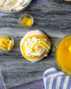 Lemon pavlova with a bowl of lemon curd and lemon zest and slices near by.