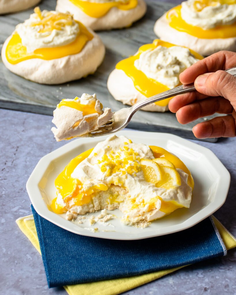 A fork lifting a piece of pavlova off a white plate with more pavlovas in the background.