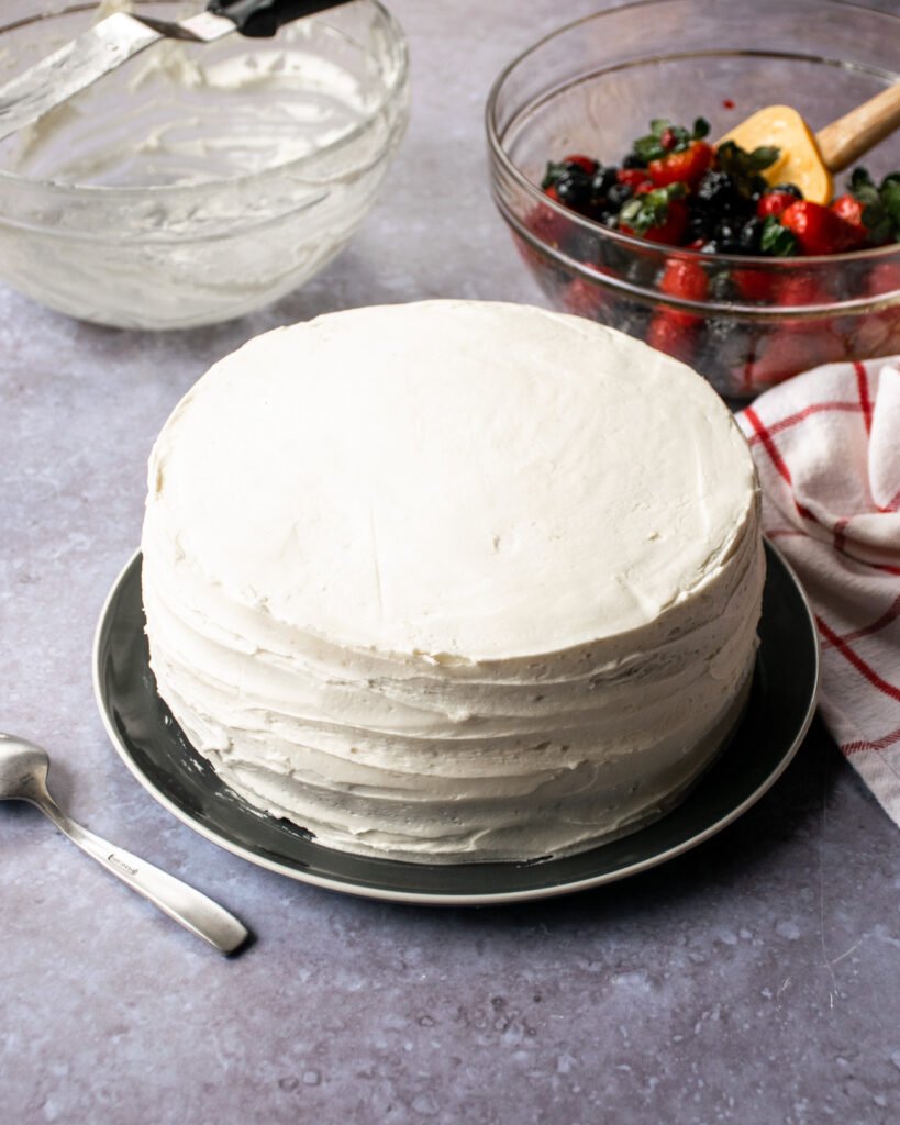 A vanilla layer cake assembled and iced on a plate with a bowl of berries near by.
