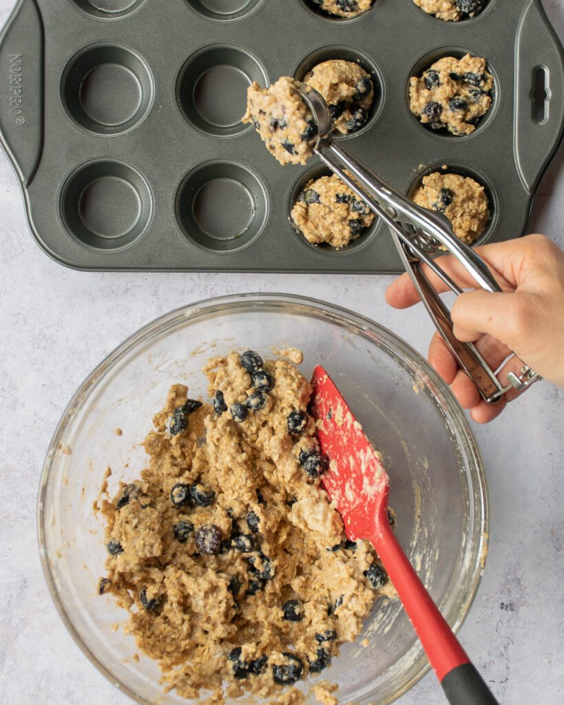 A hand scooping muffin batter into a mini muffin pan with a bowl of batter near by.