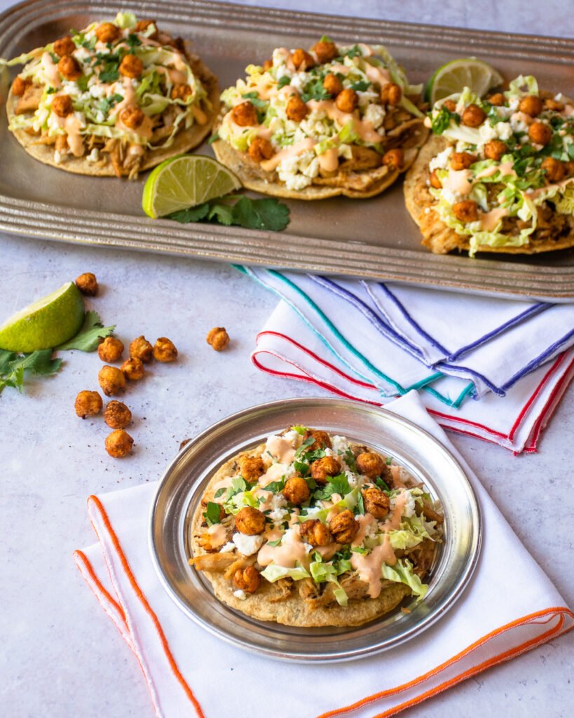 A plate with a chicken tostada in front of a silver platter with three more tostadas.